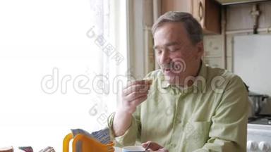 有胡子的老人端着一杯热饮和饮料。 他坐在窗边，在家吃<strong>早饭</strong>